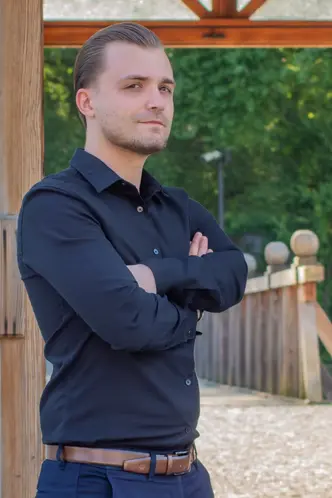 Wyprostowany mężczyzna dumnie pozuje na tle parku. Ubrany jest w cieną koszulę włożoną w spodnie.