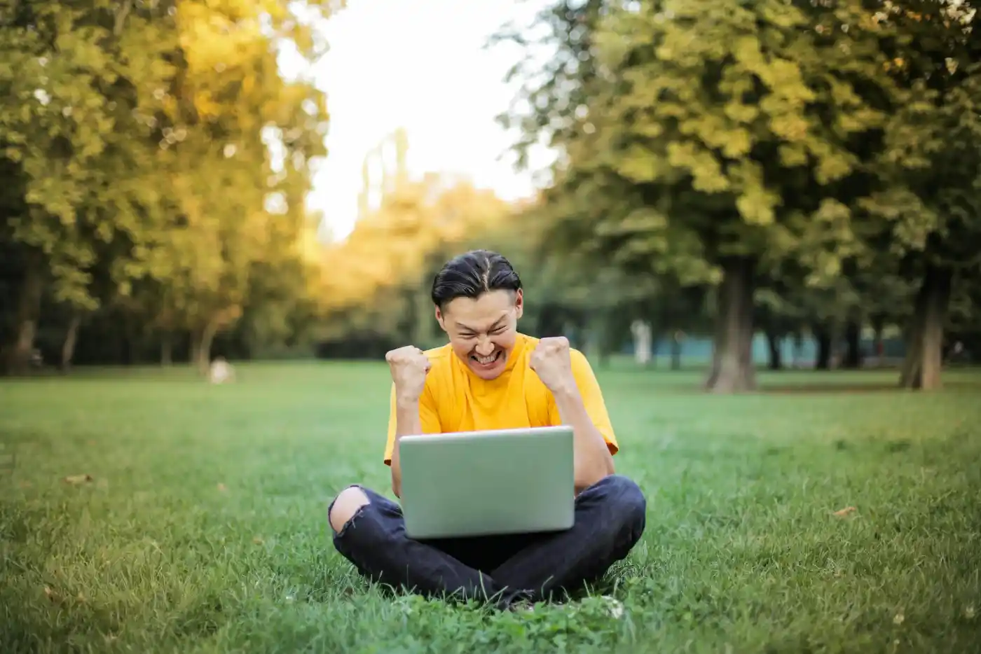 Mężczyzna z laptopem w plenerze Bardzo szczęśliwy mężczyzna siedzi "po turecku" na trawie z laptopen na kolanach, zaciska pięści w geście zwycięstwa.